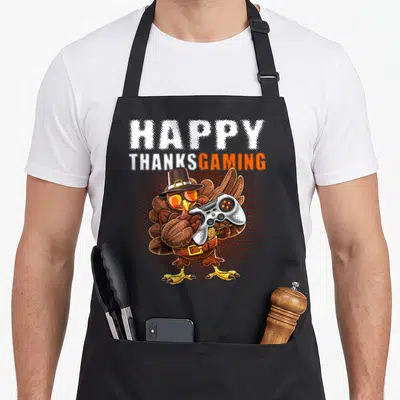Video pilgrim apron - Cartoon turkey with pilgrim hat holding game controller under Happy Thanksgaming text — shown on a black cotton canvas apron against a white background