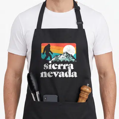 Nevada bigfoot apron - Retro landscape with Bigfoot silhouette, mountains, sun, and stacked sierra nevada text in red — shown on a black cotton canvas apron against a white background