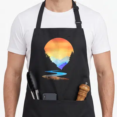 Sunset outdoor apron - Circular watercolor sunset with blue mountains, river flowing into grass, orange and yellow sky — shown on a black cotton canvas apron against a white background