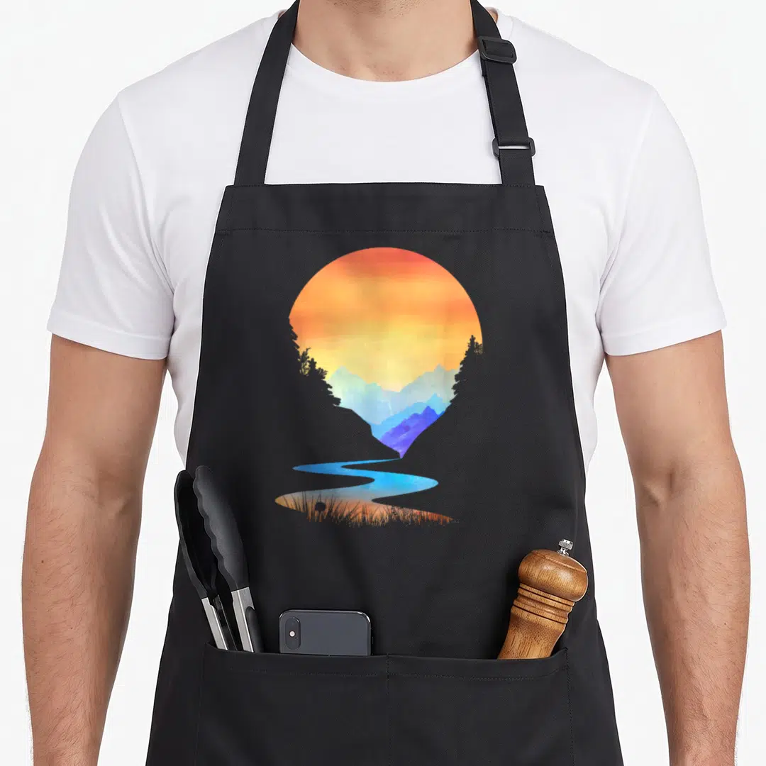Sunset outdoor apron - Circular watercolor sunset with blue mountains, river flowing into grass, orange and yellow sky — shown on a black cotton canvas apron against a white background