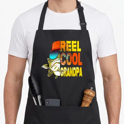 Father reel fishing apron - Cartoon fish in orange hat and blue sunglasses beside REEL COOL GRANDPA text in yellow — shown on a black cotton canvas apron against a white background