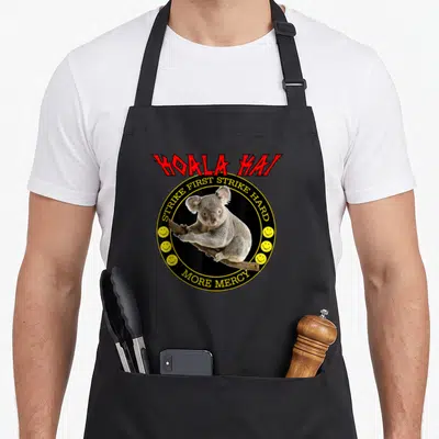 Koala art problems apron - Red KOALA KAI text above koala in black circle with yellow border and smiley faces — shown on a black cotton canvas apron against a white background