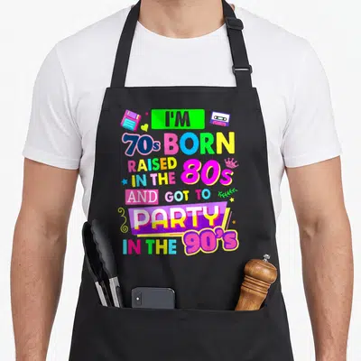 Rave 90s apron - Colorful retro text with floppy disk, cassette tape, and crown icons in neon pink, blue, yellow — shown on a black cotton canvas apron against a white background
