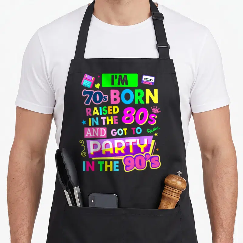 Rave 90s apron - Colorful retro text with floppy disk, cassette tape, and crown icons in neon pink, blue, yellow — shown on a black cotton canvas apron against a white background