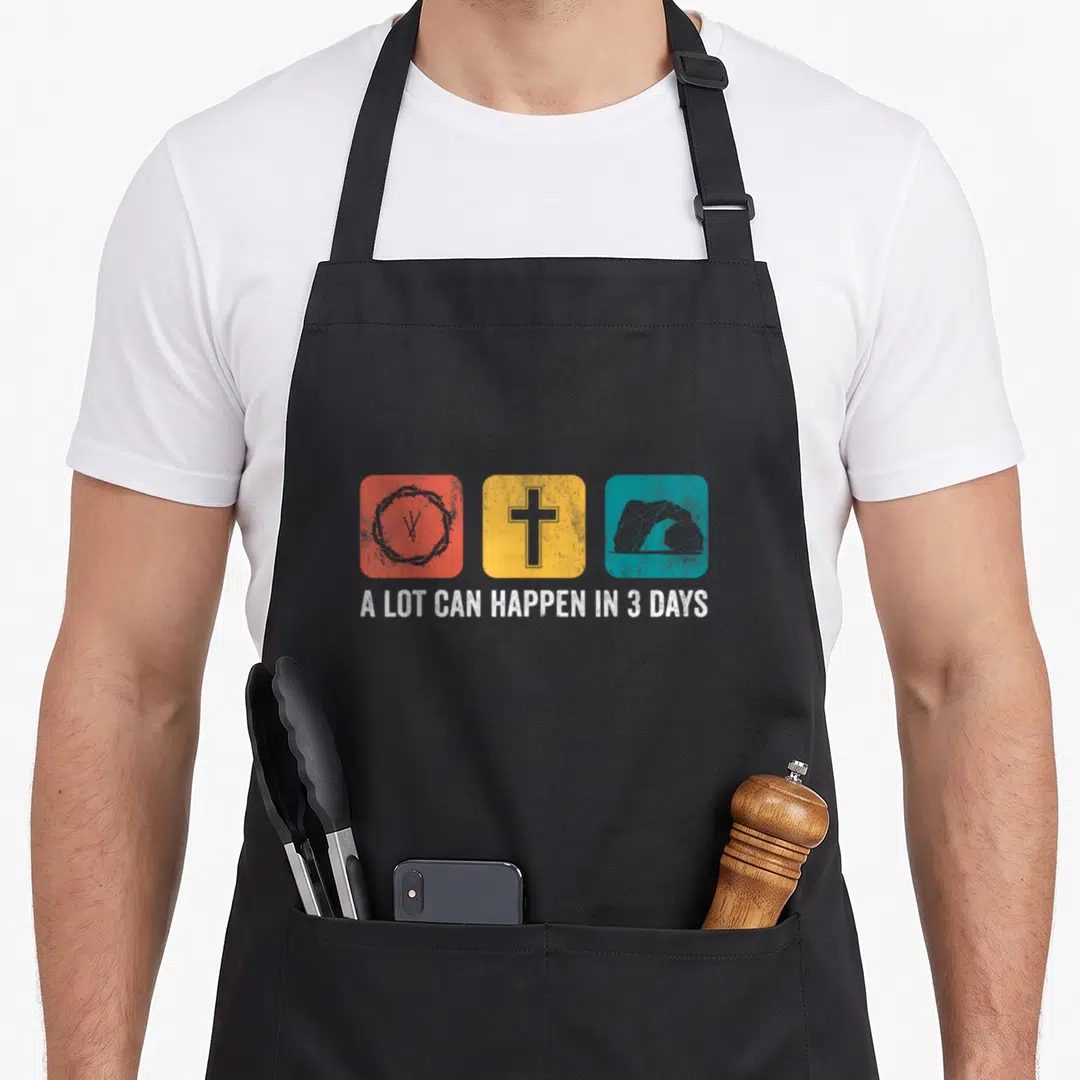 Happen easter apron - Three colored squares with crown of thorns cross and tomb above white text A Lot Can Happen in 3 — shown on a black cotton canvas apron against a white background