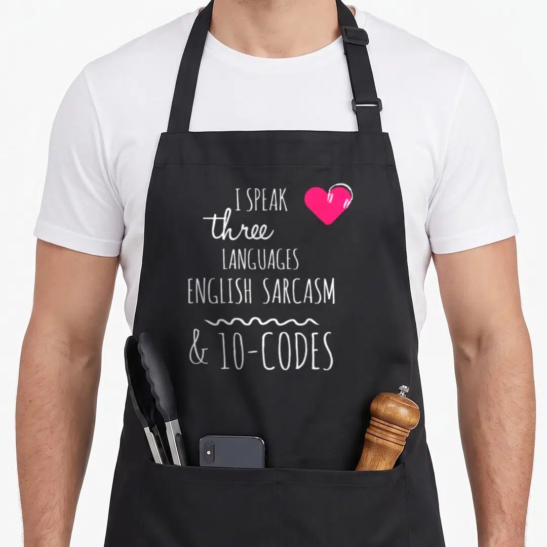 Sarcasm dispatcher apron - White text I speak three languages English sarcasm and 10-codes next to pink heart — shown on a black cotton canvas apron against a white background