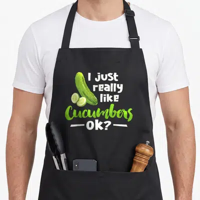 Cucumber veggie apron - Cartoon cucumber and two slices beside I just really like Cucumbers Ok text — shown on a black cotton canvas apron against a white background