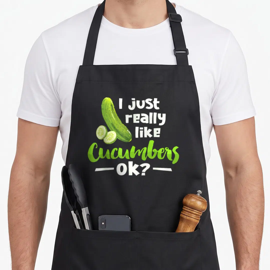 Cucumber veggie apron - Cartoon cucumber and two slices beside I just really like Cucumbers Ok text — shown on a black cotton canvas apron against a white background