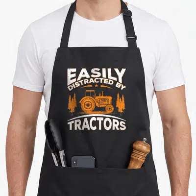 Easily farming apron - White text EASILY DISTRACTED BY TRACTORS framing orange tractor and three trees — shown on a black cotton canvas apron against a white background