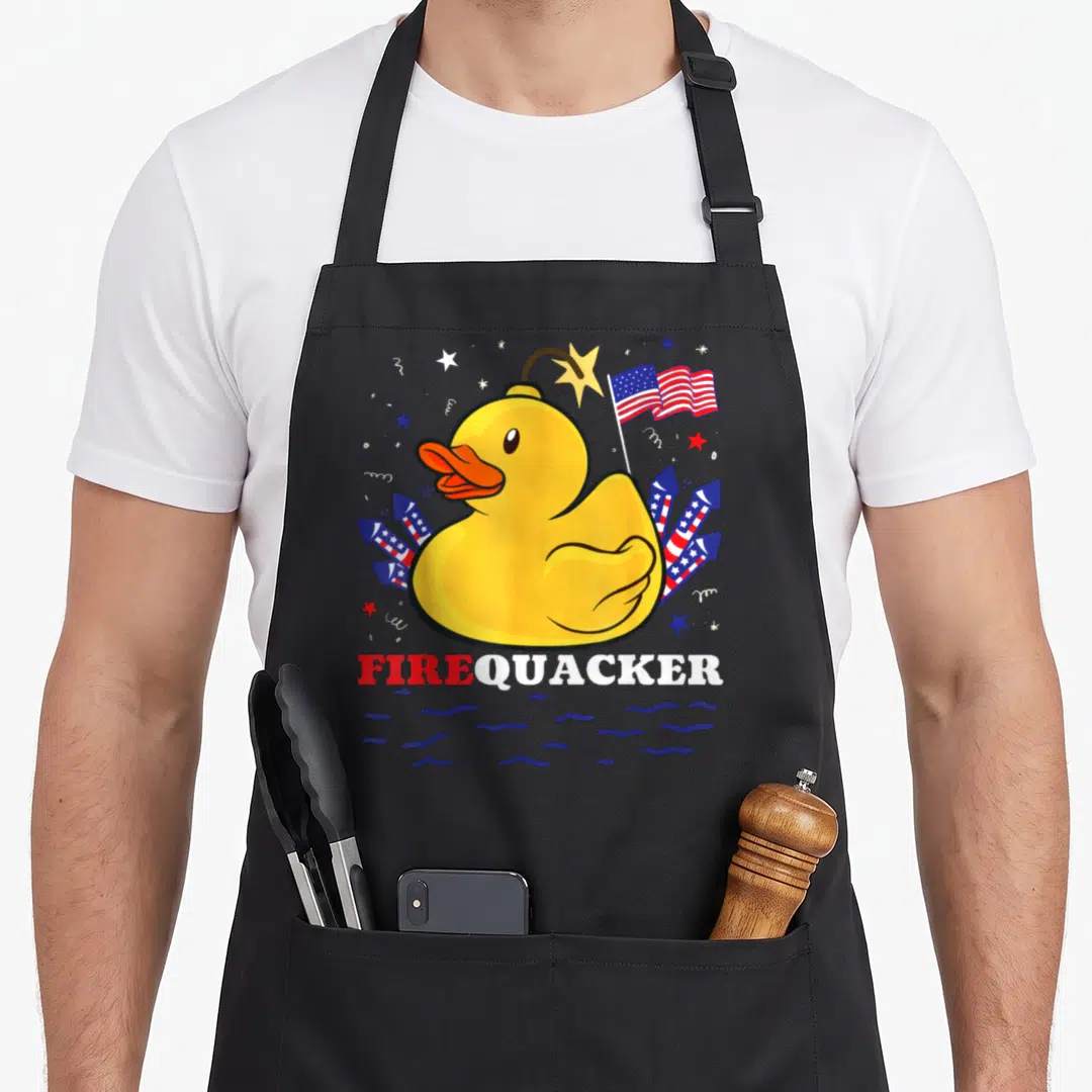 Firecracker duck 4th of july apron - Yellow rubber duck with USA flag and FIREQUACKER text over blue water — shown on a black cotton canvas apron against a white background