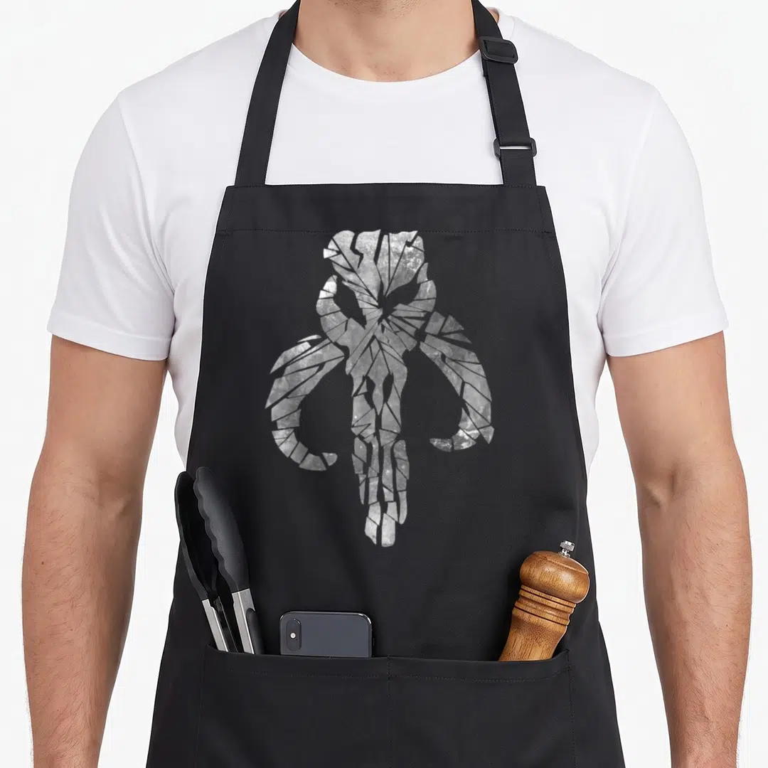 Clan skull apron - White cracked skull with horns on black background, distressed texture — shown on a black cotton canvas apron against a white background