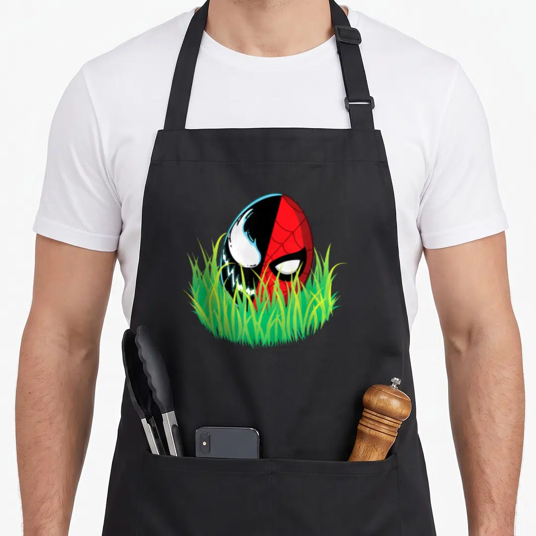 Vintage easter egg apron - Cartoon egg split vertically into red and black character faces hiding in green grass — shown on a black cotton canvas apron against a white background