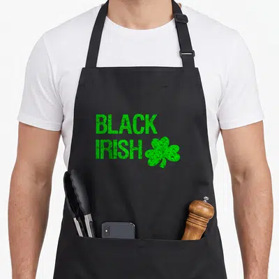 Bright st patrick day aprons - Distressed green BLACK IRISH text with shamrock on black background — shown on a black cotton canvas apron against a white background