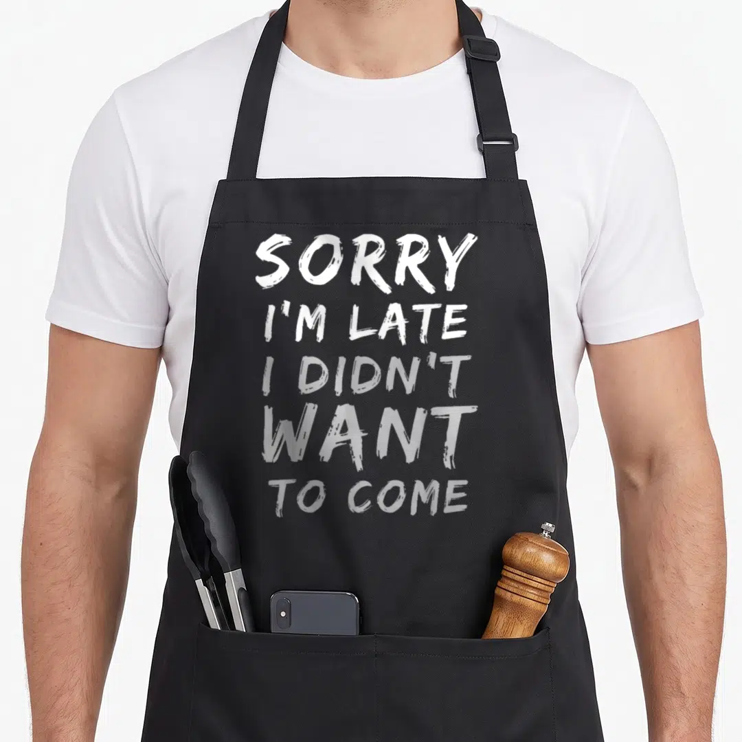 Come late apron - Stacked white and grey brush-stroke text reading sorry i'm late i didn't want to come — shown on a black cotton canvas apron against a white background