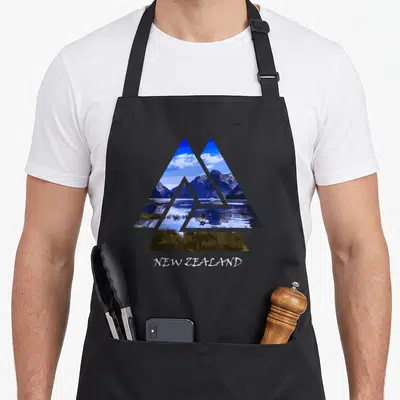 Milford new zealand apron - Geographic triangle containing blue mountain lake scene with white text NEW ZEALAND below — shown on a black cotton canvas apron against a white background