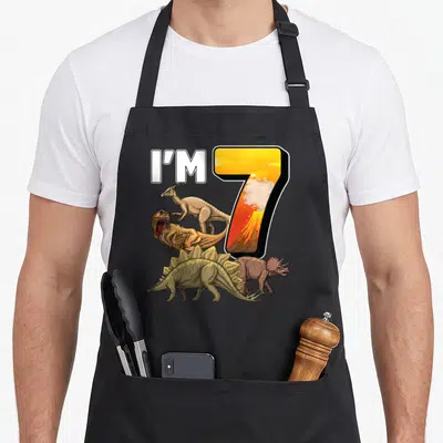Birthday 7th dinosaur apron - I'M 7 text with four dinosaurs and volcano inside number seven in yellow and brown — shown on a black cotton canvas apron against a white background