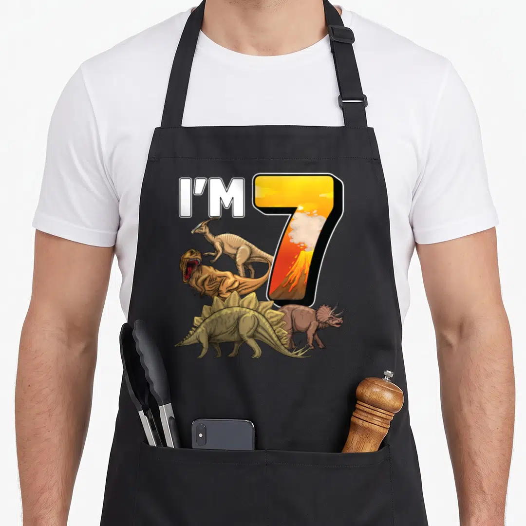 Birthday 7th dinosaur apron - I'M 7 text with four dinosaurs and volcano inside number seven in yellow and brown — shown on a black cotton canvas apron against a white background