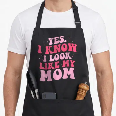 Cute mother daughter apron - Stacked pink text reading Yes I Know I Look Like My Mom surrounded by small stars on black — shown on a black cotton canvas apron against a white background