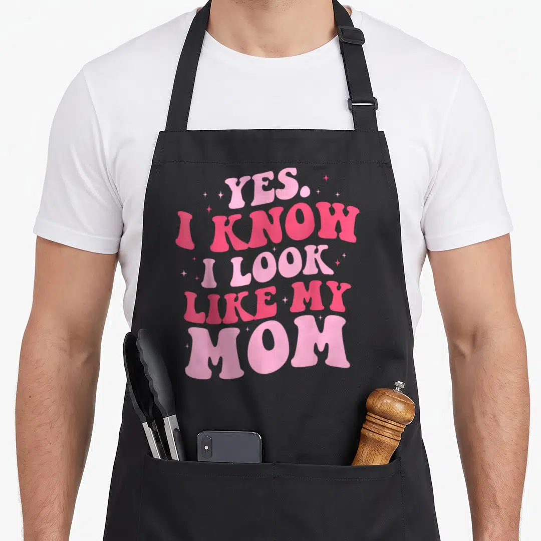Cute mother daughter apron - Stacked pink text reading Yes I Know I Look Like My Mom surrounded by small stars on black — shown on a black cotton canvas apron against a white background