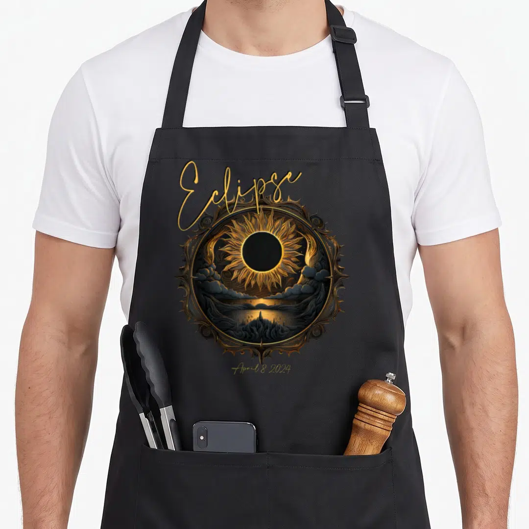 Totality april apron - Ornate circular frame with solar eclipse, mountains, water, clouds, and arched text reading — shown on a black cotton canvas apron against a white background