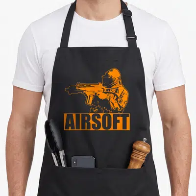 Airsoft combat apron - Orange cartoon airsoft player holding rifle above AIRSOFT text block — shown on a black cotton canvas apron against a white background