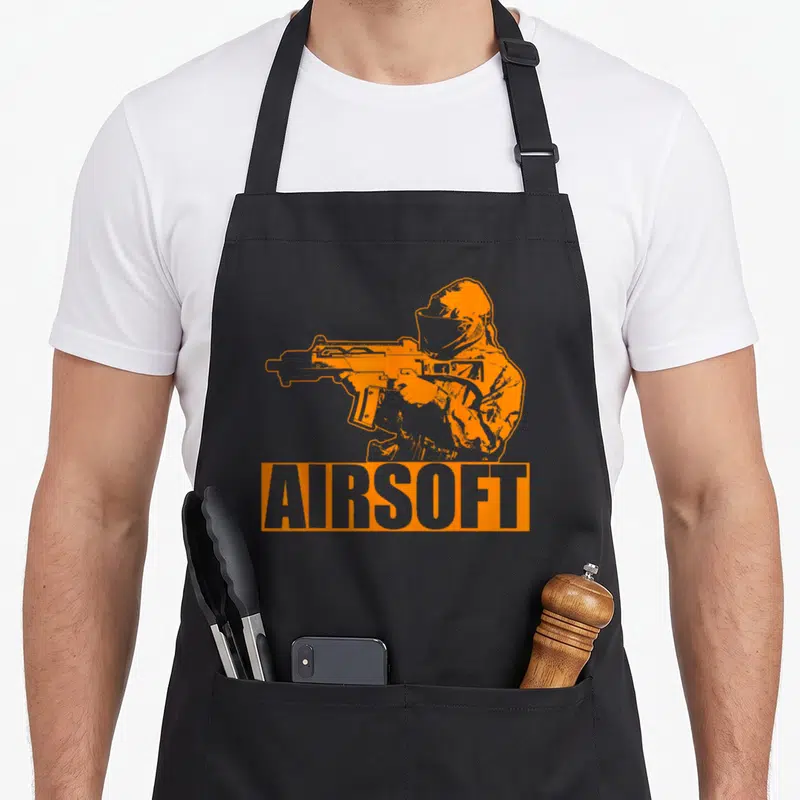 Airsoft combat apron - Orange cartoon airsoft player holding rifle above AIRSOFT text block — shown on a black cotton canvas apron against a white background