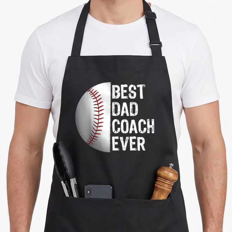 Coach baseball apron - Left half of a white baseball with red stitching beside stacked white distressed text — shown on a black cotton canvas apron against a white background
