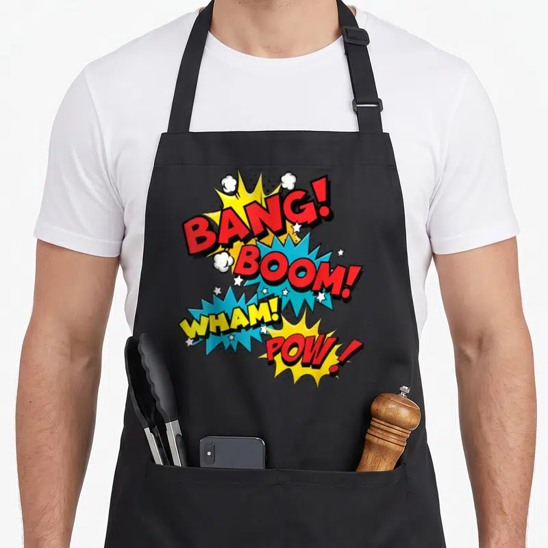 Bang pop art apron - Four red yellow blue and white comic bursts with BANG BOOM WHAM POW text and stars — shown on a black cotton canvas apron against a white background