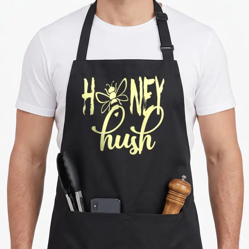 Honey hush lover apron - Pale yellow bee inside HONEY word above script hush text — shown on a black cotton canvas apron against a white background