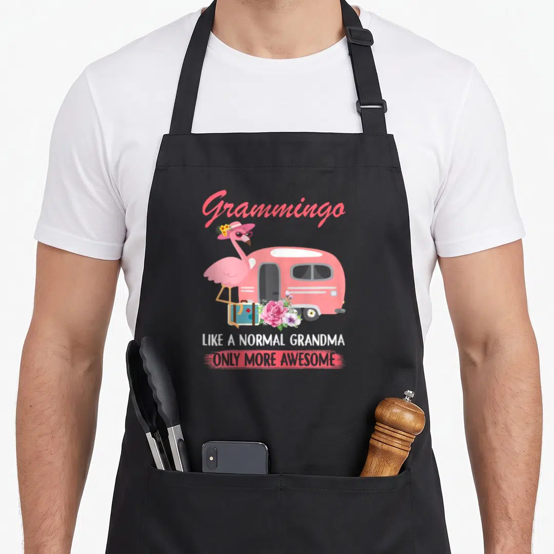 Grammingo normal grandma apron - Pink flamingo standing by camper van with suitcase and flowers under Grammingo text — shown on a black cotton canvas apron against a white background