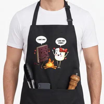 Smore camp chef apron - Cartoon s'more characters with I Love You S More speech bubbles and hearts over a campfire — shown on a black cotton canvas apron against a white background