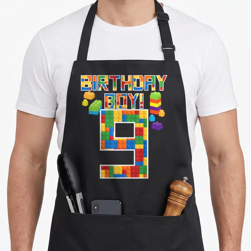 9th building aprons - BIRTHDAY BOY! 9 text and number filled with colorful toy bricks and scattered blocks — shown on a black cotton canvas apron against a white background