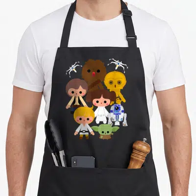 Funny heroe kawaii apron - Chibi Star Wars characters and two X-wings in brown, gold, white, blue, green, and red — shown on a black cotton canvas apron against a white background