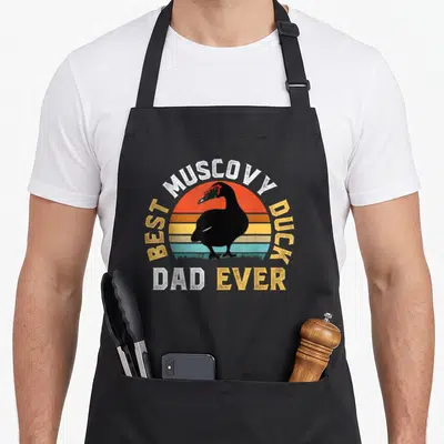 Muscovy cotton duck apron - Black muscovy duck silhouette over striped orange yellow teal sun with text Best Muscovy — shown on a black cotton canvas apron against a white background