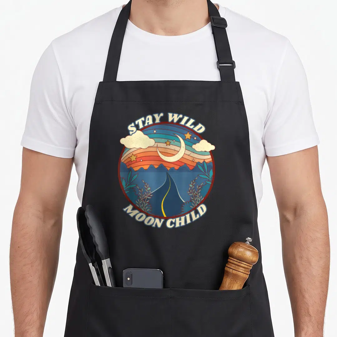 Stay boho apron - Retro circular design with STAY WILD MOON CHILD text, road, mountains, and crescent moon in blue — shown on a black cotton canvas apron against a white background