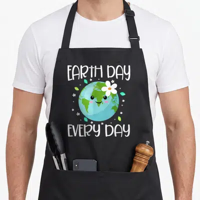 Student everyday apron - Cartoon Earth with face and flower, EARTH DAY EVERY DAY text, leaves, stars — shown on a black cotton canvas apron against a white background