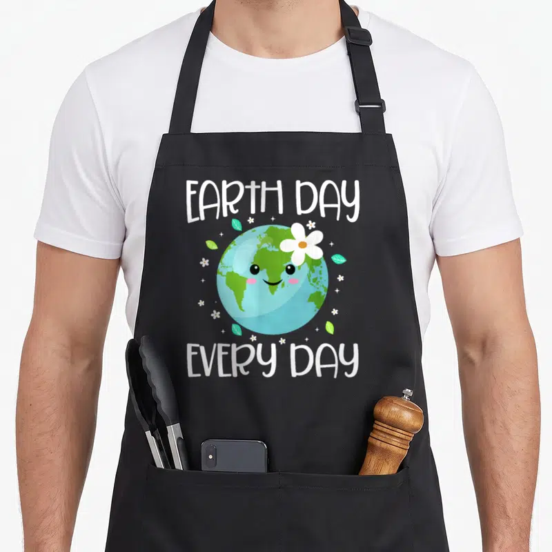 Student everyday apron - Cartoon Earth with face and flower, EARTH DAY EVERY DAY text, leaves, stars — shown on a black cotton canvas apron against a white background