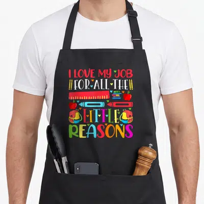 Vintage reason teacher apron - Stacked text I love my job with ruler, books, apples, backpacks and hearts in red blue — shown on a black cotton canvas apron against a white background