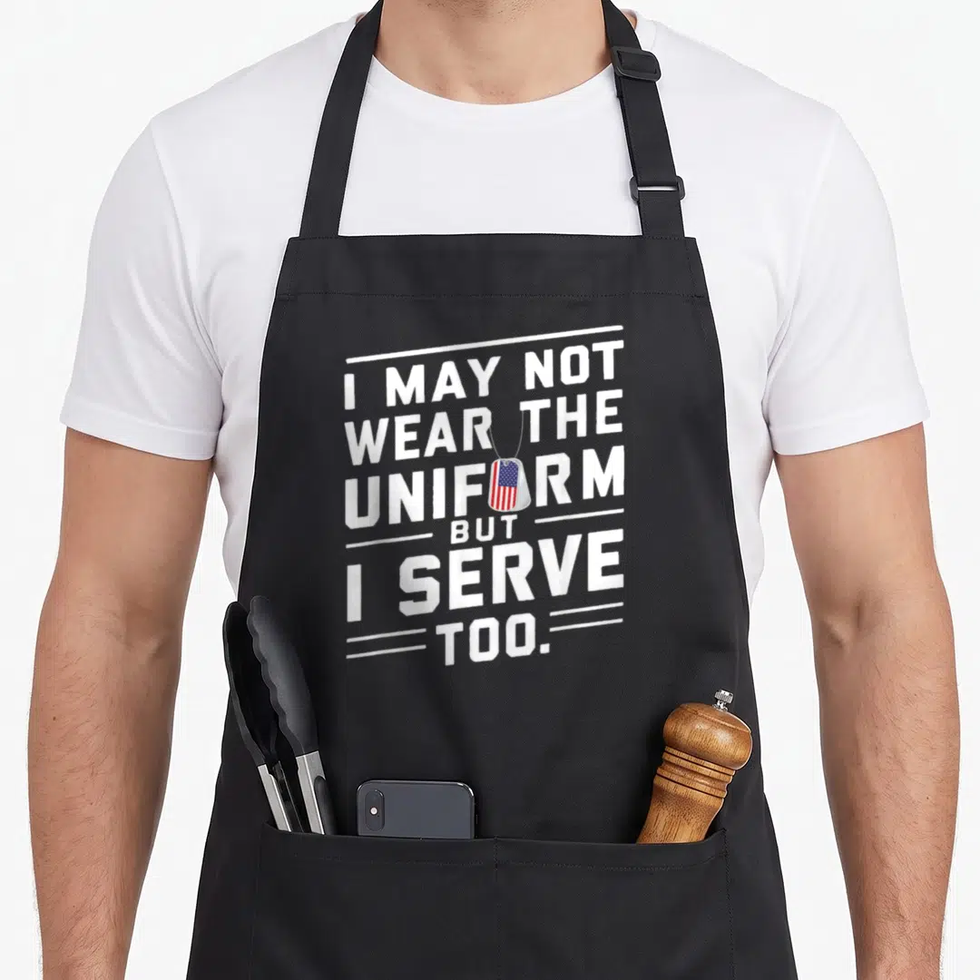 Flag personalized dog apron - Stacked white text reading I may not wear the uniform but I serve too with a flag dog tag — shown on a black cotton canvas apron against a white background