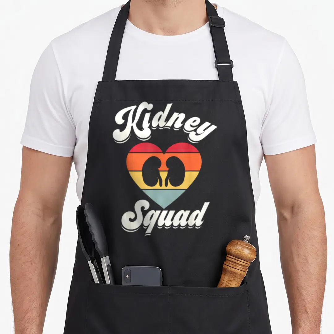 Kidney nurse apron - Retro heart with two kidney silhouettes and Kidney Squad text in white, red, orange, yellow — shown on a black cotton canvas apron against a white background