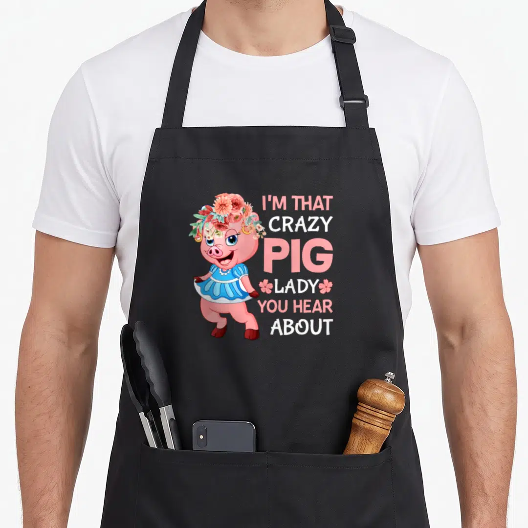 Hear crazy pig lady apron - Cartoon pink pig in blue dress and flower crown beside stacked text reading I'm That Crazy — shown on a black cotton canvas apron against a white background