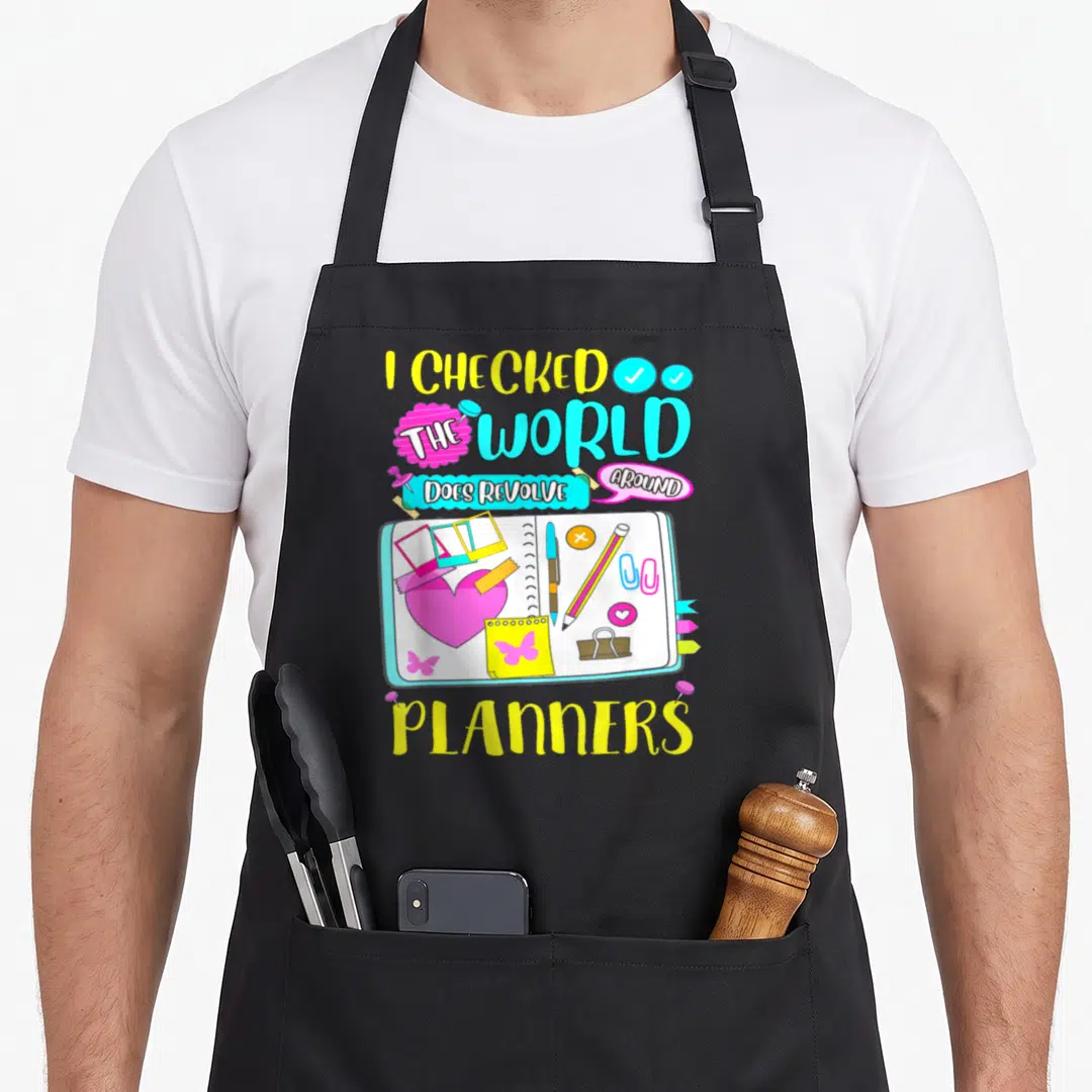 Doe creative addict apron - Open planner with heart, pens, flags, and text I checked the world does revolve around — shown on a black cotton canvas apron against a white background