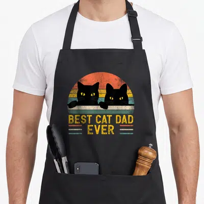 Creature cat apron - Two black cats peeking over grey ledge with BEST CAT DAD EVER text on retro striped background — shown on a black cotton canvas apron against a white background