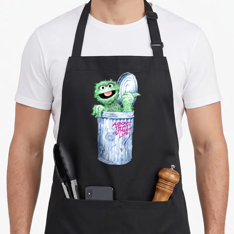 Funny oscar the grouch apron - Green monster in blue trash can with pink About That Street Life text — shown on a black cotton canvas apron against a white background
