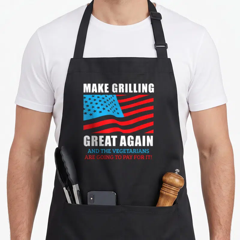 Trump master apron - Make Grilling Great Again text over red white blue USA flag with vegetarian joke — shown on a black cotton canvas apron against a white background