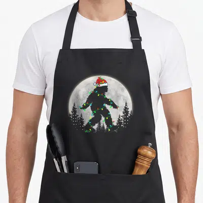 Sasquatch christmas tree apron - Grey bigfoot silhouette in red santa hat wrapped in multicolor lights against full moon — shown on a black cotton canvas apron against a white background