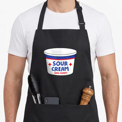 Sour cream aprons - White sour cream tub with blue text, two red stars, and 100% YUMMY slogan — shown on a black cotton canvas apron against a white background