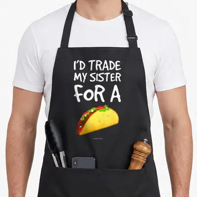 Trade taco apron - I'D TRADE MY SISTER FOR A text above colorful cartoon taco illustration — shown on a black cotton canvas apron against a white background