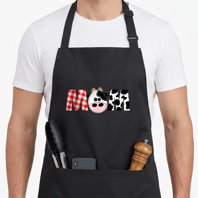Birthday farm 1st cow print apron - MOM text with red gingham M, cartoon cow O, and black spotted M — shown on a black cotton canvas apron against a white background