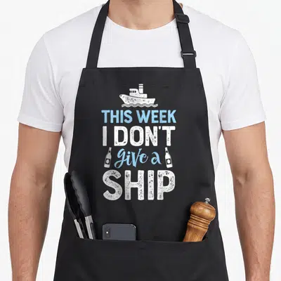 Week trips apron - White ship and two bottles flanking distressed text THIS WEEK I DON'T give a SHIP — shown on a black cotton canvas apron against a white background